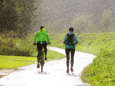 Hier sehen Sie zwei Personen draußen im Regen sport machen. Die eine Person fährt Rad, die ander Person joggt daneben. 