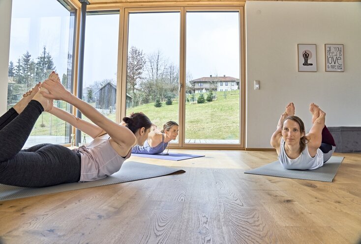 Drei Damen liegen für eine Yoga-Übung mit dem Bauch auf je einer Matte und ziehen ihre Füße mit den Händen über den Rücken. Durch die großen Fenster im Hintergrund sind eine grüne Wiese mit Bäumen und der blaue Himmel zu sehen.
