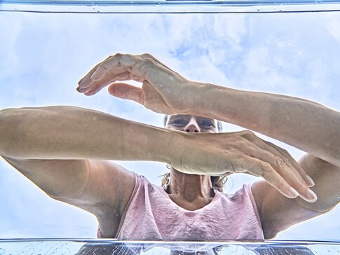 Hier sehen Sie eine Frau mit pinkem Shirt, welche ihre Arme in klares Wasser taucht. Die Aufnahme wurde aus dem Wasser, unterhalb der Frau, aufgenommen.