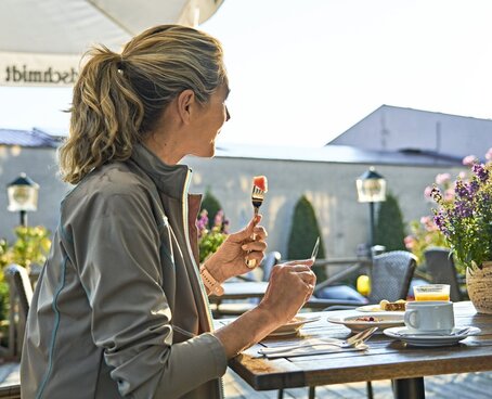 Hier sehen Sie eine Frau mit blondem Haar auf der Terrasse sitzen, welche gesundes Obst geniesst.