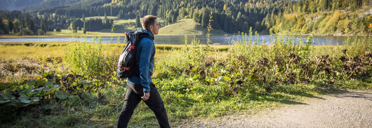 Hier sehen Sie den ehemaligen Profi-Fußballer Philipp Lahm. In Wanderbekleidung geht er auf einem Schotterweg spazieren. Hinter ihm sind Berge und ein See zu sehen. 