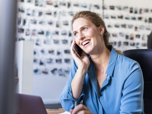 Hier sehen Sie eine Frau in Businesskleidung an einem Schreibtisch sitzen. Sie telefoniert mit einem Handy und lacht. Hinter ihr ist eine Wand zu sehen, an welcher Fotos hängen. 