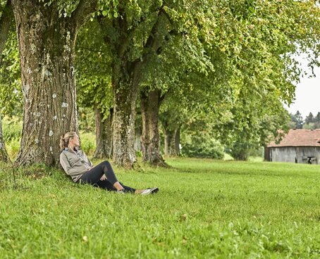 Eine Frau sitzt auf einer Wiese im Gras und lehnt sich an einem großen Baum an. Im Hintergrund sind viele Bäume und eine kleine Hütte zu sehen.