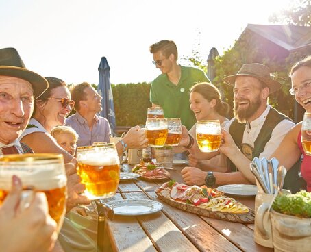 Sie sehen 7 Personen an einem Tisch im Biergarten sitzen, mit Bier in der Hand und einem Brotzeitbrettl auf dem Tisch.