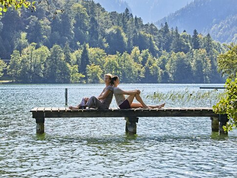 Hier sehen Sie einen Steg von der Seite der in einen großen See läuft. Auf dem Steg sitzen eine Frau und ein Mann, Rücken an Rücken und genießen das schöne Wetter. Hinter ihnen sind Berge und Wald zu sehen. 