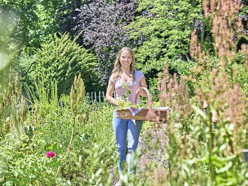 Hier sehen Sie eine junge Frau, die mit einem Korb voller Kräuter durch einen Kräutergarten spaziert.