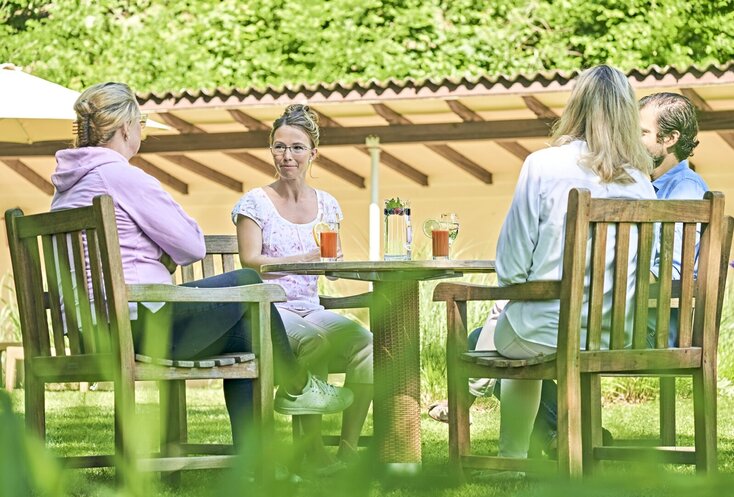Sie sehen vier Personen in einem Garten auf Holzstühlen um einen Holztisch sitzen.