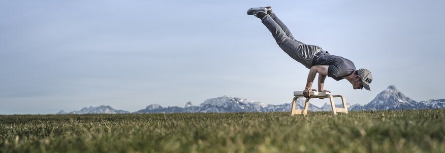 Hier sehen Sie den Bayern Botschafter Kilian Trenkle. Auf einer Wiese macht er Yogaübungen mit einem Yogahocker. Hinter ihm ist eine klassische bayerische Landschaft zu sehen.
