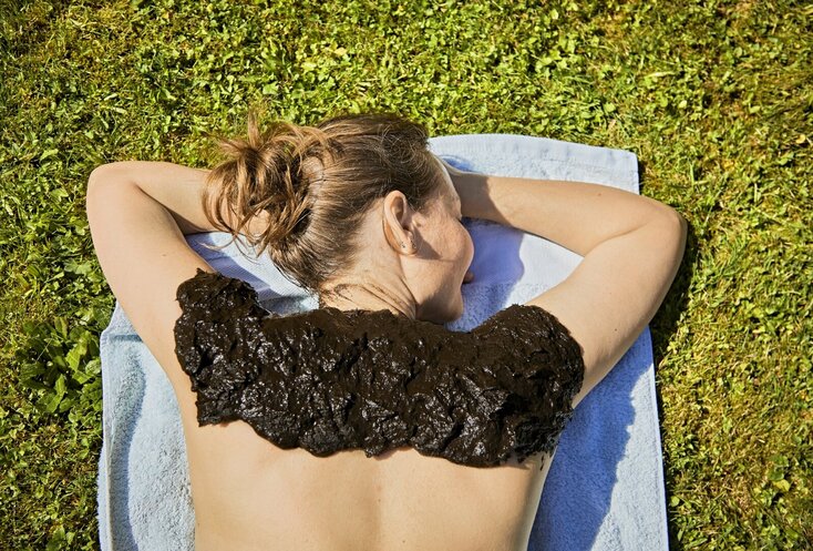 Hier sehen Sie eine Frau die auf dem Bauch mit einem Handtuch auf einer Wiese liegt. Auf ihren Schultern und Nacken wurde Moor verteilt. Sie hat ihre Augen geschlossen und genießt die angenheme Wärme.