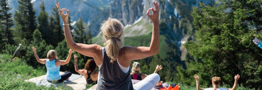Hier sehen Sie eine Gruppe von Personen, welche in den Bergen Yoga praktizieren. Alle schauen in Richtung Berge und sind mit dem Rücken zu uns gedreht. 