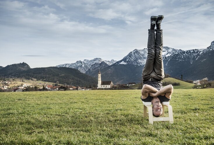 Hier sehen Sie den Bayern Botschafter Kilian Trenkle. Auf einer Wiese macht er einen Kopfstand auf einem Yogahocker. Hinter ihm ist eine klassische bayerische Landschaft zu sehen.