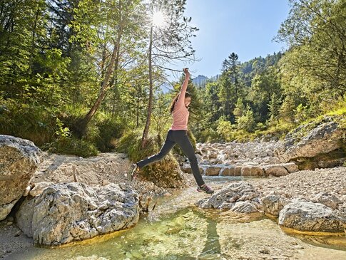 Hier sehen Sie einen kleinen Flusslauf in Mitten von Bäumen und Büschen. Eine Frau springt über diesen Flusslauf von einem Stein zum anderen Stein.