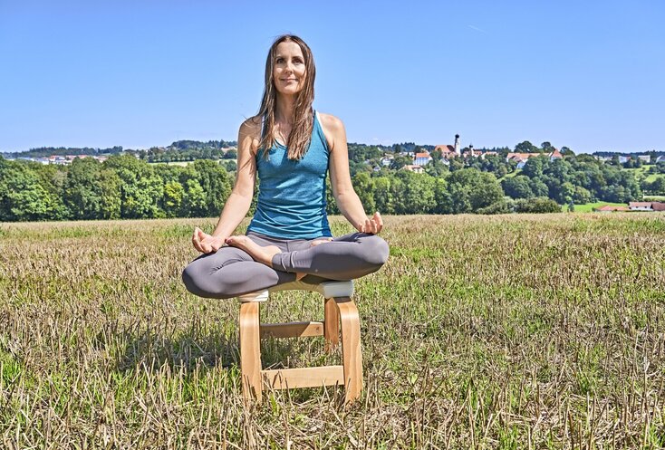 Hier sehen Sie eine junge Frau, die im Schneidersitz auf einem Holzhocker in einem Feld sitzt und meditiert.
