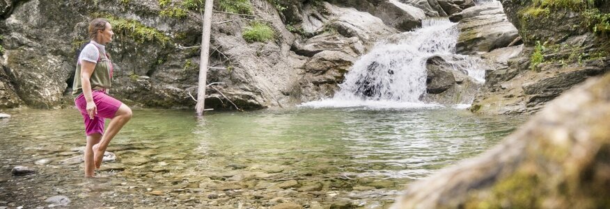 Hier sehen Sie einen kleinen Bergfluss. Bayern Botschafterin Christine Waibel-Beer steht an einer seichten Stelle im Wasser und kneippt im Storchenschritt. 