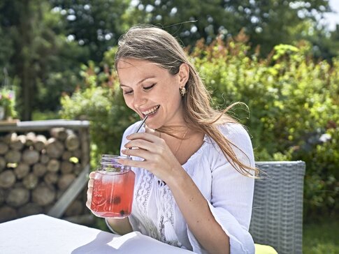 Hier sehen Sie eine junge Frau, die lächelnd draußen im Garten sitzt und Fruchtsaft trinkt. 