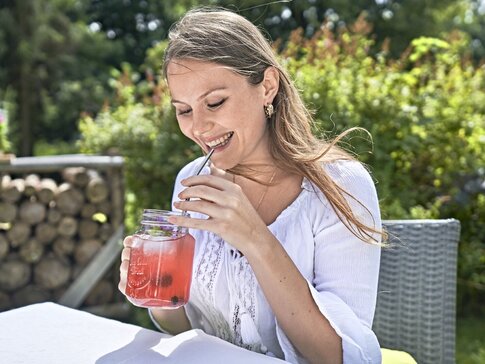 Hier sehen Sie eine junge Frau, die lächelnd draußen im Garten sitzt und Fruchtsaft trinkt. 