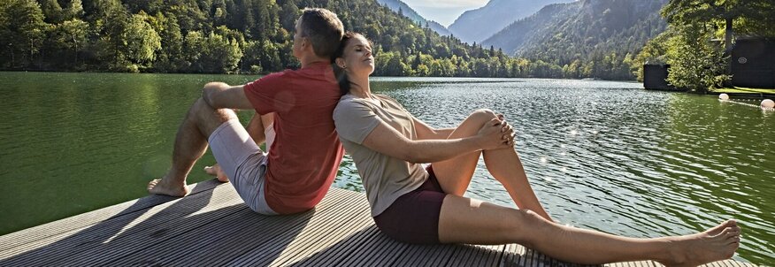Sie sehen einen Mann und eine Frau mit dem Rücken aneinandergelehnt auf einem Steg am See. Im Hintergrund sehen Sie Berge.