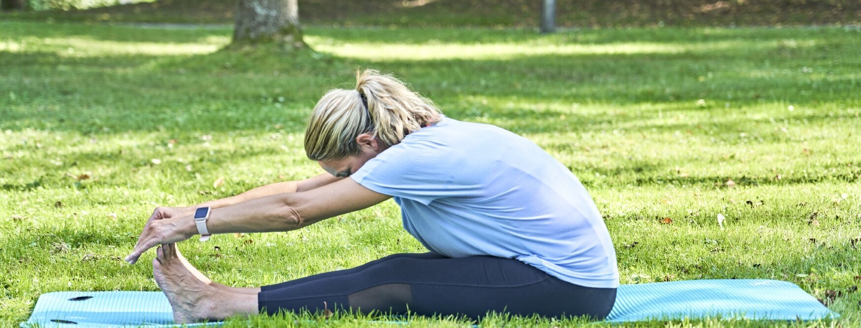 Ich sehe eine Frau mit schwarzer Sporthose und hellblauem T-shirt, die sich auf einer türkisen Yoga-Matte dehnt. 