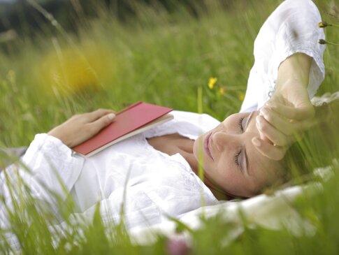 Hier sehen Sie eine Frau in einer Wiese liegen. Sie hat ihre Augen geschlossen, auf ihrem Bauch hat sie in Buch liegen. 