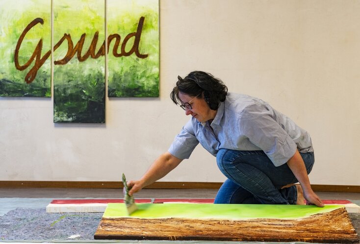 Hier sehen Sie die Expertin Monika Aigner, wie sie auf dem Boden ein Bild auf eine Leinwand malt.
