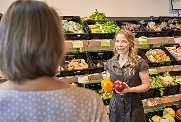 Sie sehen Ernährungsberaterin Sarah Reinmold in einem dunklen Kleid in der Obst- und Gemüseabteilung eines Supermarkts stehen. Sie steht vor den mit Gemüse und Obst gefüllten Regalen und hält eine rote und eine gelbe Paprikaschote in den Händen. Im Vordergrund ist eine Frau mit halblangem, dunkelblondem Haar und einer hellen Bluse mit Punkten von hinten zu sehen.