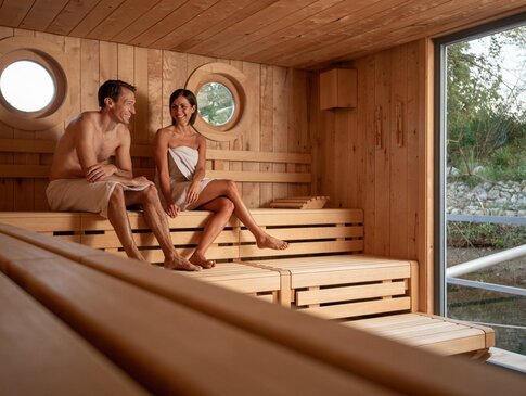 Hier sehen Sie zwei Personen in einer Sauna sitzen. Beide haben ein Handtuch umgewickelt. Die Sauna hat ein Fenster welches Blick nach draußen gewährt.