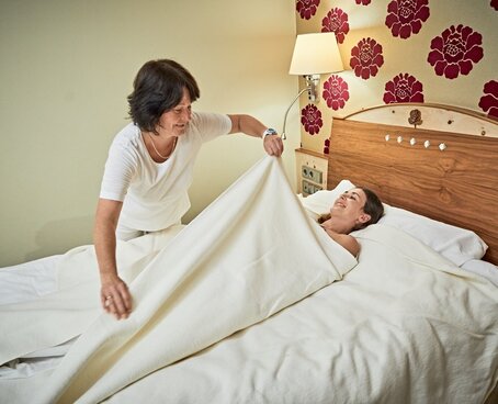 Hier sehen Sie eine Frau die von einer Therapeutin im Bett mit Decken und Tüchern komplett eingewickelt wird. Nur der Kopf bleibt frei.