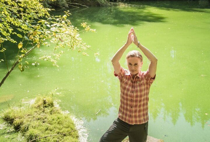Hier sehen Sie die Bayern Botschafterin und Waldbademeisterin Elke Seidel. Sie steht auf einem Felsen am Wasser und macht die Yoga Postition "der Baum".