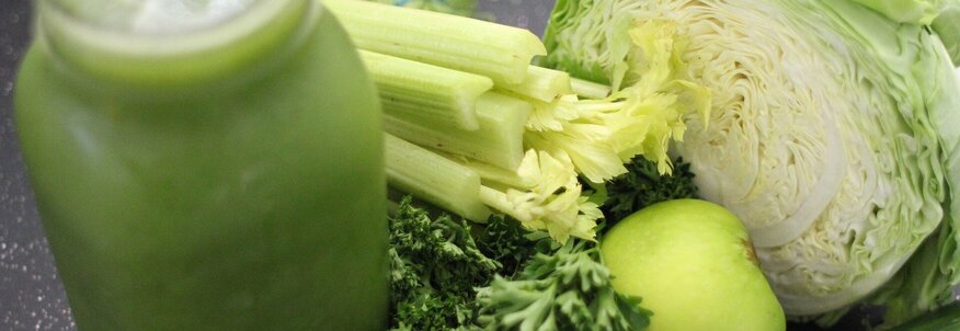 Hier sehen Sie verschiedene grüne Gemüse und Obst Sorten, welche zur zubereitung für einen grünen Smoothie notwendig sind. Neben dem Gemüse steht auch ein Glas, gefüllt mit Smoothie.