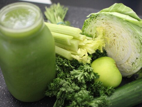 Hier sehen Sie verschiedene grüne Gemüse und Obst Sorten, welche zur zubereitung für einen grünen Smoothie notwendig sind. Neben dem Gemüse steht auch ein Glas, gefüllt mit Smoothie.