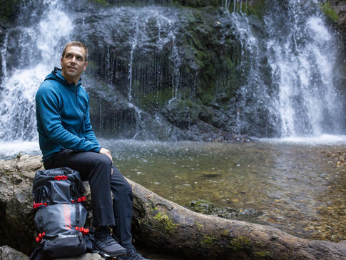 Hier sehen Sie den ehemaligen Fußballprofi Philipp Lahm. Er sitzt au einem Holzstamm in der Nähe eines Wasserfalls. Er hat den Rucksack neben sich abgelegt und macht Rast. Hinter ihm sieht man den Wasserfall und das seichte Gewässer. 