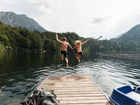 Eine Frau und ein Mann springen händehaltend von einem Steg in einen See. Neben dem Steg befindet sich ein Tretboot. Der See ist umgeben von Wald und Bergen.