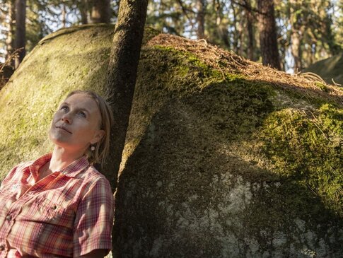 Hier sehen Sie eine Dame, welche sich an einem moosigen Stein im Wald anlehnt. Sie blickt in den Himmel rauf.