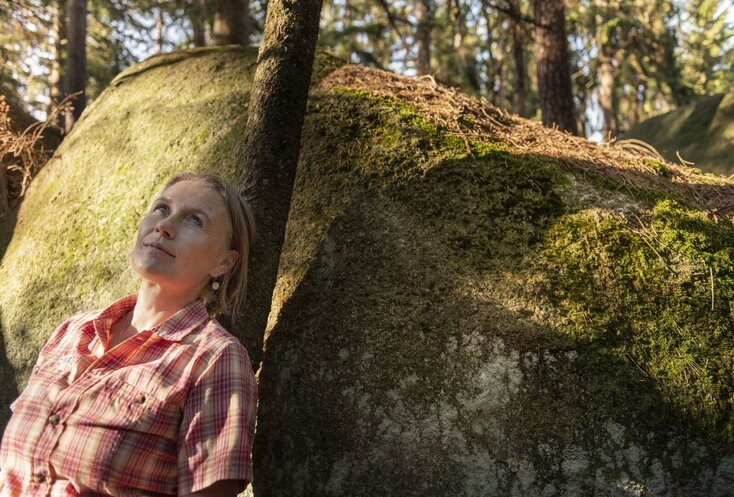 Hier sehen Sie eine Dame, welche sich an einem moosigen Stein im Wald anlehnt. Sie blickt in den Himmel rauf.