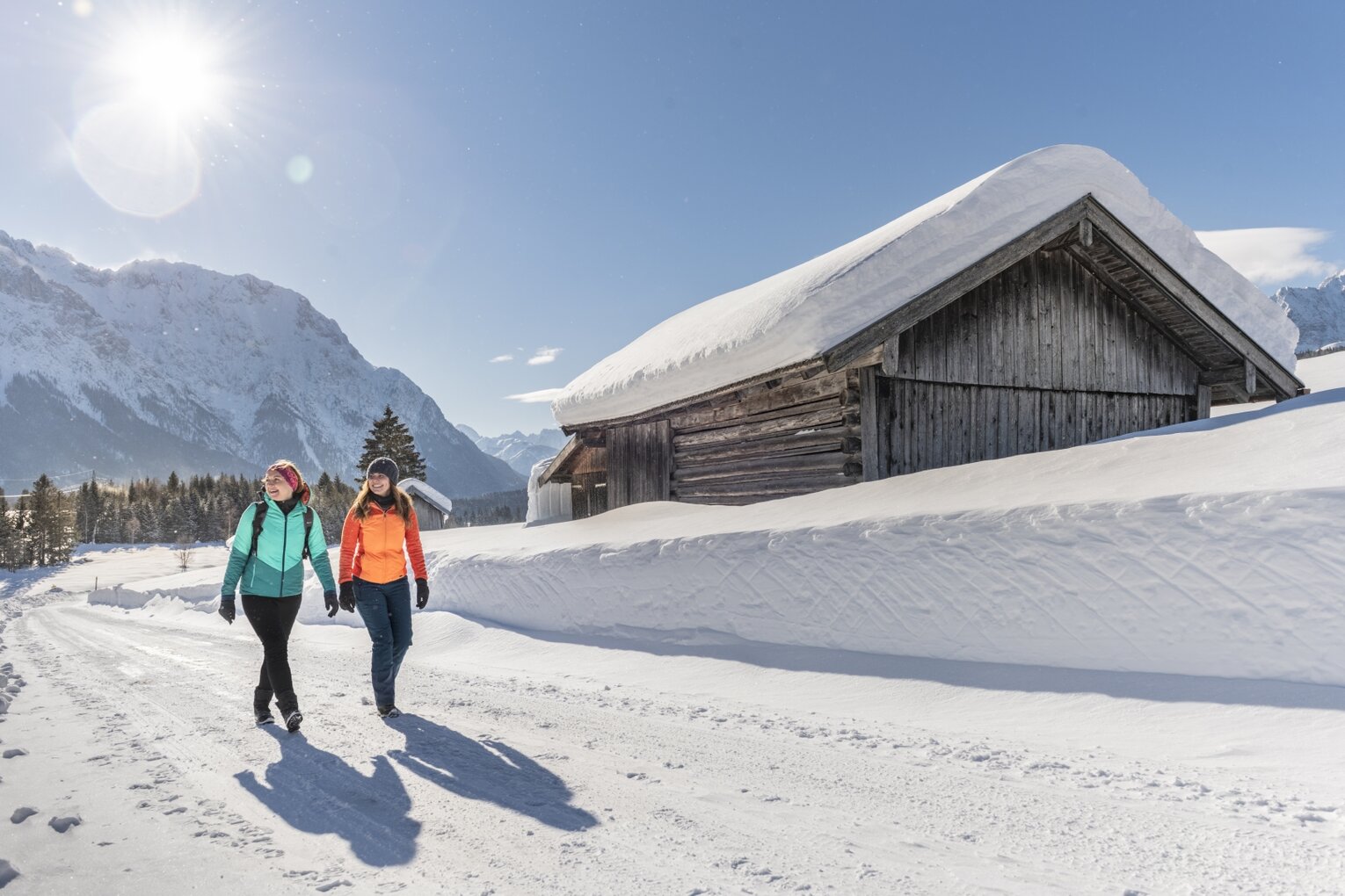 Hier sehen Sie zwei Personen in einer Winterlandschaft wandern. Sie gehen auf einer geräumten Straße, neben ihnen ist der Schnee noch sehr hoch. Sie gehen an einer kleinen Hütte vorbei und die Sonne scheint. 
