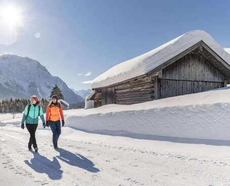 Hier sehen Sie zwei Personen in einer Winterlandschaft wandern. Sie gehen auf einer geräumten Straße, neben ihnen ist der Schnee noch sehr hoch. Sie gehen an einer kleinen Hütte vorbei und die Sonne scheint. 