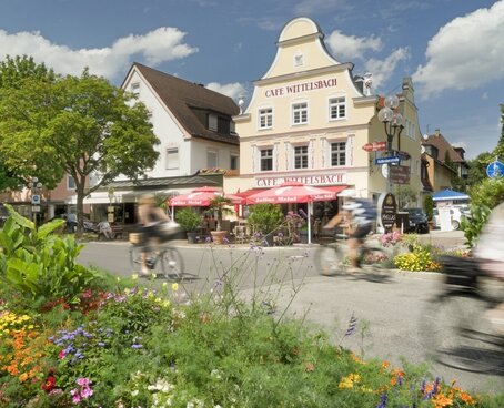 Sie sehen die Innenstadt von Bad Wörishofen. Im Vordergrund sind Blumen in Beeten zu sehen, dahinter eine gepflasterte Straße und Gebäude mit Sonnenschirmen und Tischen und Stühlen davor. Menschen fahren mit dem Rad vorbei.
