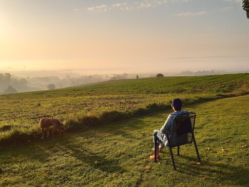 Hier sehen Sie eine Frau von hinten, die auf einem Camping-Stuhl sitzt und den sonnig-dunstigen Ausblick über Felder und hinunter in ein Tal genießt.