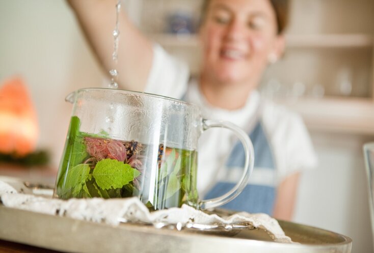 Hier sehen Sie eine Teekanne aus Glas in der viele frische Kräuter sind. Eine Frau im Hintergrund ist unscharf zu sehen. Sie schüttet heißes Wasser in die Teekanne und brüht den Tee auf. 