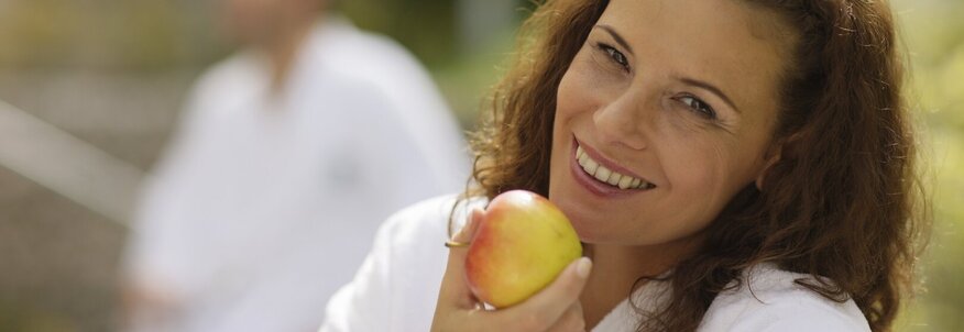 Hier sehen Sie eine Frau in einem Bademantel, sie lacht in die Kamera und hat einen Apfel in der Hand. 