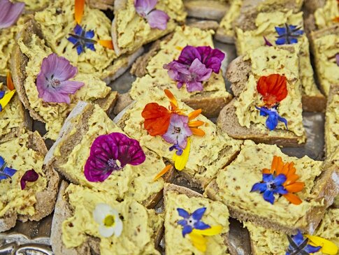 Hier sehen Sie einige Scheiben dunkles Bauernbrot. Bestrichen ist das Brot mit einem gelben curry Aufstrich. Dekoriert ist jedes Brot mit einer essbaren Blüte.