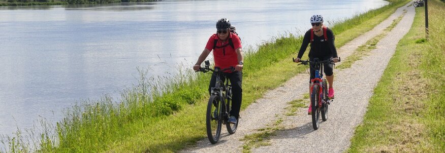 Hier sehen Sie zwei Personen auf einem Radweg radfahren. Der Weg führt entlang an einem Fluss. 