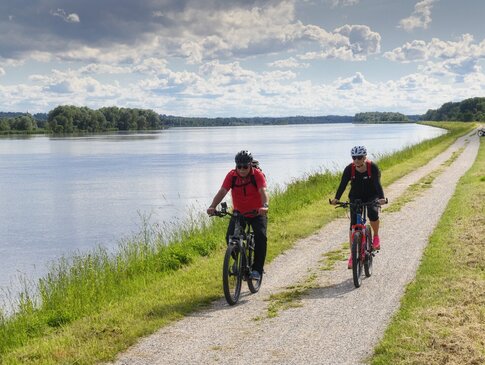 Hier sehen Sie zwei Personen auf einem Radweg radfahren. Der Weg führt entlang an einem Fluss. 