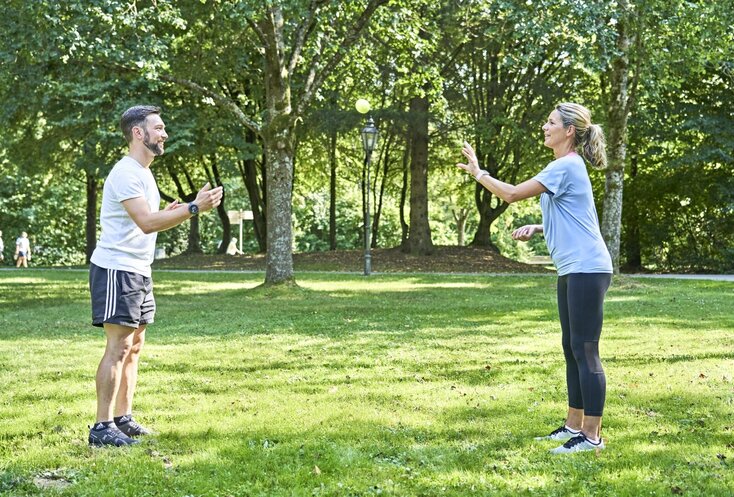 Ein Mann und eine Frau stehen sich in einem Park in Sportoutfit gegenüber. Der Mann wirft mit der rechten Hand einen Tennisball, den die Frau mit rechts auffangen will.