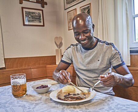 Hier sehen Sie Michel Frenzel-Assih lächelnd an einem Tisch sitzen. Vor ihm steht ein Rinderbraten mit Kloß und Blaukraut und ein Bier.