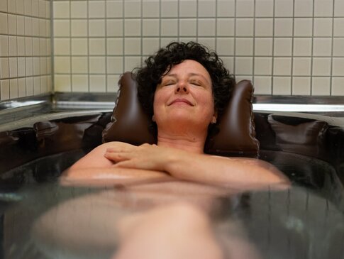 Hier sehen Sie eine Frau in einr Stahlbadewanne liegen. Ihr Kopf stützt sich auf einem Nackenkissen ab. 