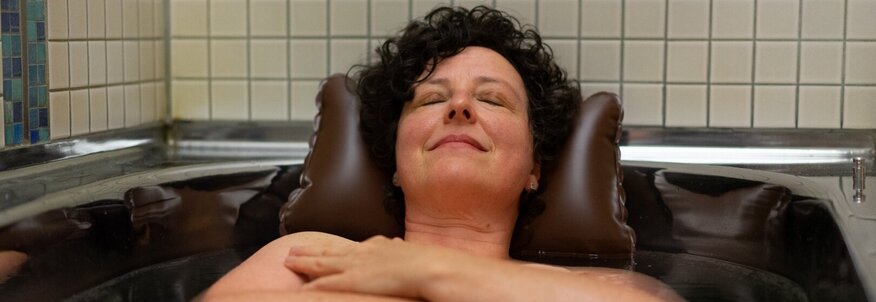 Hier sehen Sie eine Frau in einr Stahlbadewanne liegen. Ihr Kopf stützt sich auf einem Nackenkissen ab. 