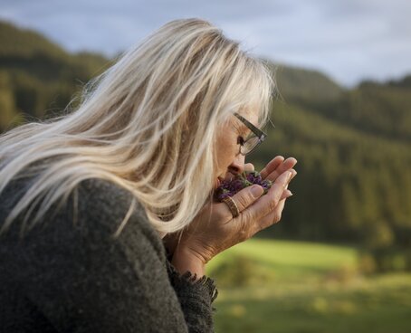 Hier sehen Sie eine Frau die draußen in der Natur an frisch geschnittenen Kräutern und Blumen in ihrer Hand riecht. Dabei hat sie ihre Augen geschlossen. Hinter ihr sind Wiesen und Wälder. 