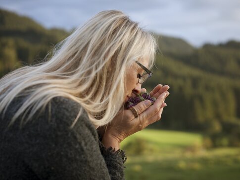 Hier sehen Sie eine Frau die draußen in der Natur an frisch geschnittenen Kräutern und Blumen in ihrer Hand riecht. Dabei hat sie ihre Augen geschlossen. Hinter ihr sind Wiesen und Wälder. 