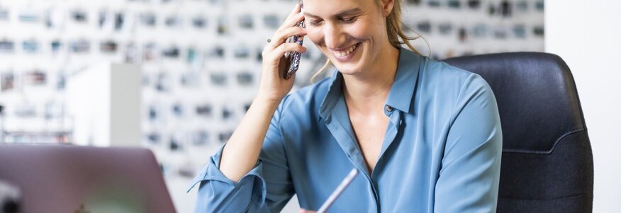 Ich sehe eine braunhaarige Frau, die eine blaue Bluse trägt. Sie sitzt grinsend vor ihrem Laptop, hat einen weißen Stift in der Hand und telefoniert. 